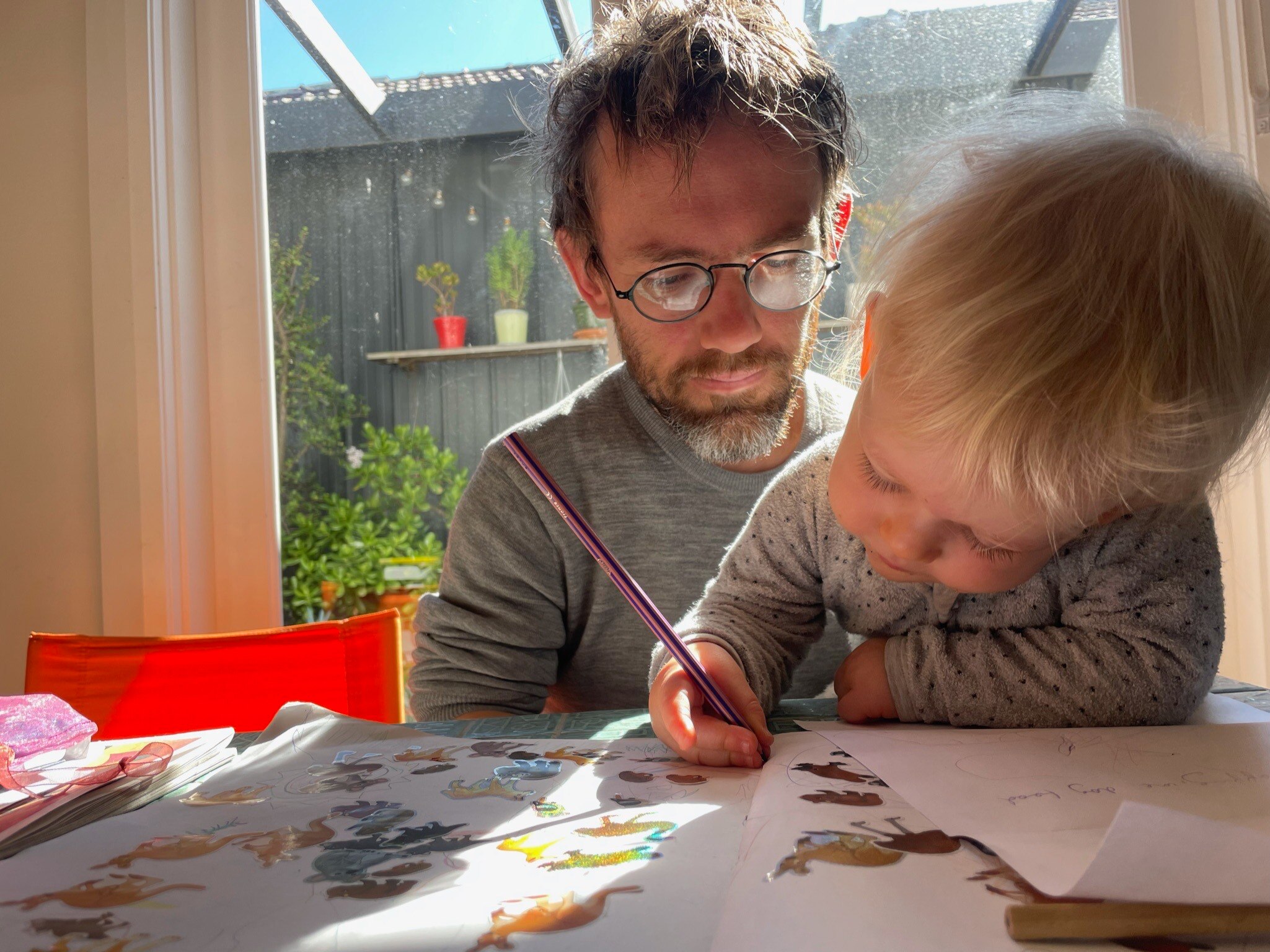 A man in glasses looks over the shoulder of a young girl who sits on his lap, drawing at the table.