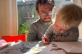 A man in glasses looks over the shoulder of a young girl who sits on his lap, drawing at the table.