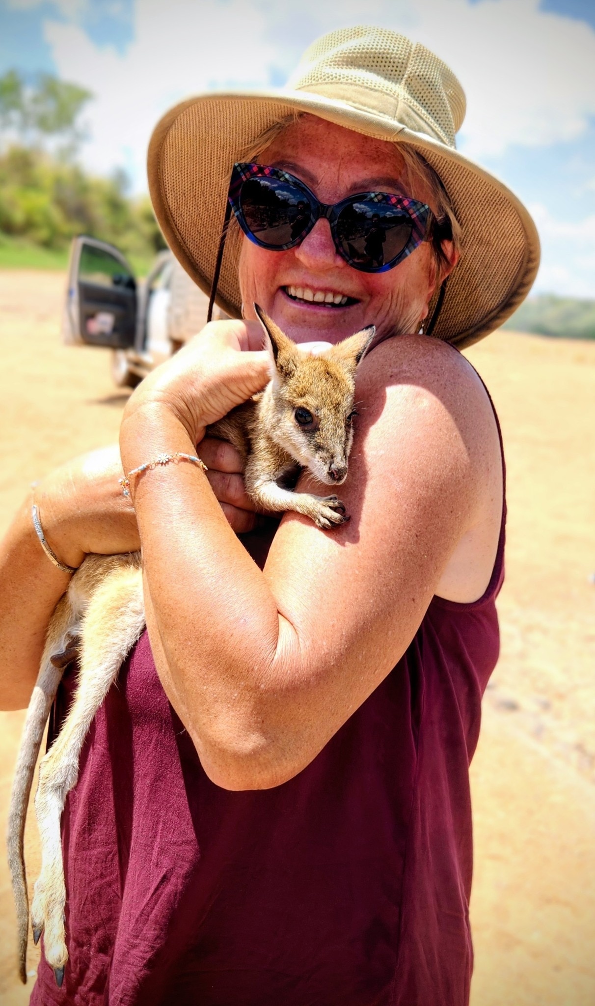 Pensioner Heather Janine wearing a hat and sunglasses, smiling and holding a baby kangaroo.