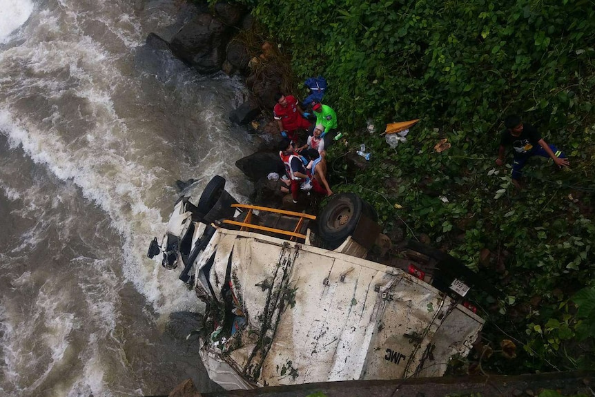 A heavily damaged, white truck lies on its side on a riverbank with rescuers helping to remove the driver