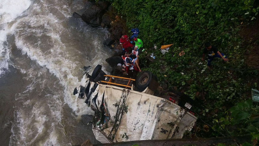 A heavily damaged, white truck lies on its side on a riverbank with rescuers helping to remove the driver