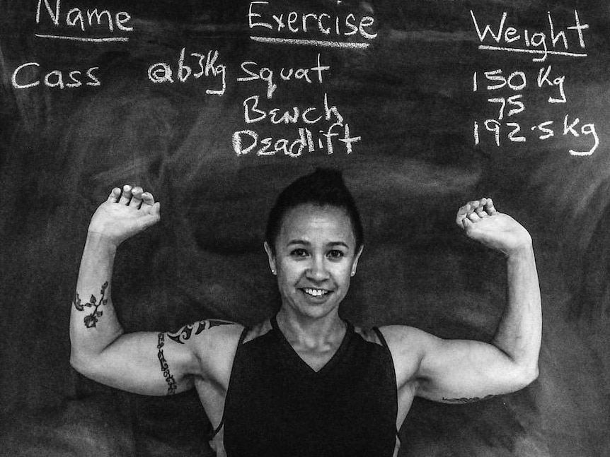 Cass Pickard stands in front of a blackboard displaying her personal bests - 150kg squat, 75kg bench, 192.5kg deadlift