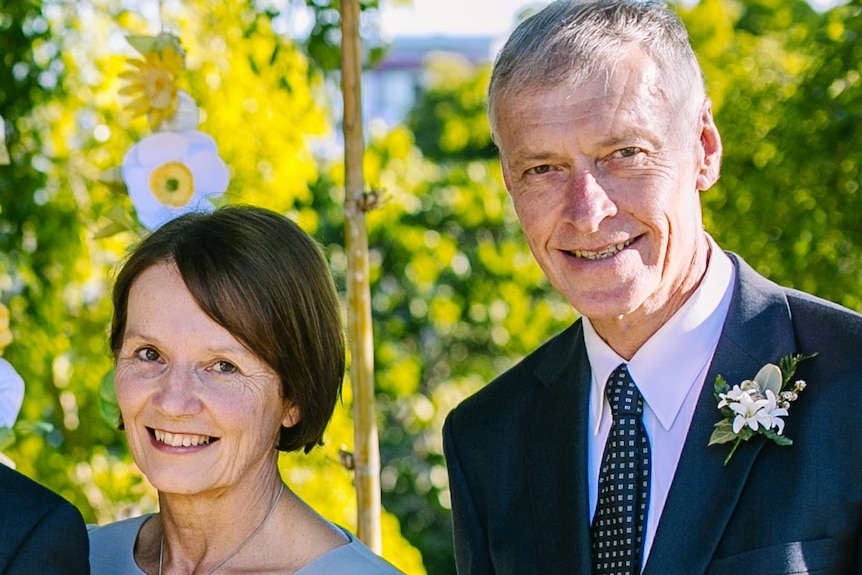 Jill and Roger Guard smile, wearing formalwear as they pose for a photo in a sunlit garden setting.