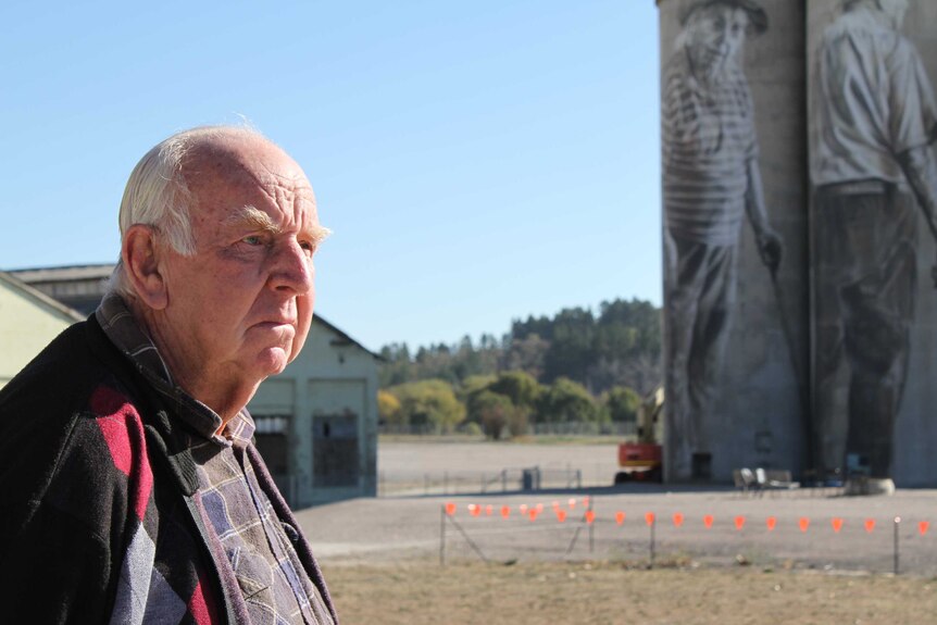 An older man with white hair standing near silos with artwork painted on them.