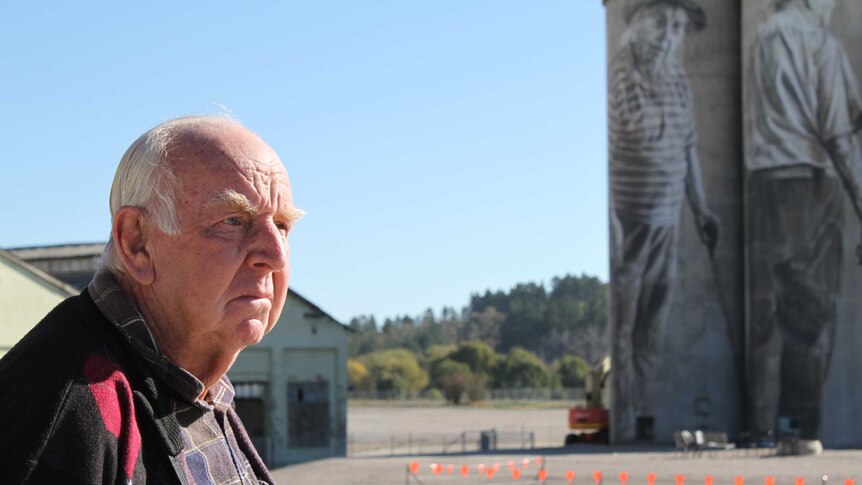 An older man with white hair standing near silos with artwork painted on them.