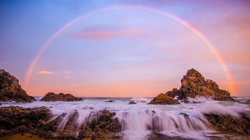 A full rainbow forms over the ocean at dusk, with rocks in the foreground.