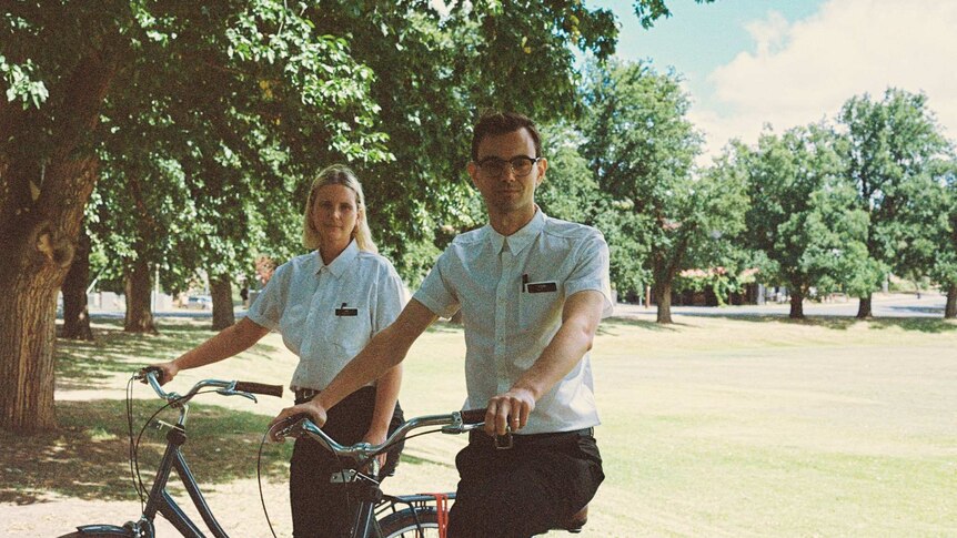 Jo and Tom dressed in crisp, white buttoned up short-sleeved shirts and black slacks, sitting on bikes, in a sunny park