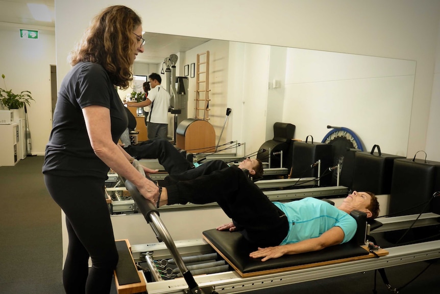 A woman lies on an exercise machine while a female instructor stretches her leg.