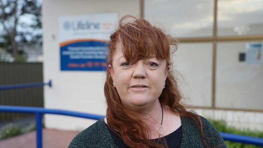 A woman with long orange hair and a green shirt stands in front of a while building with blue railings