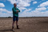 A man in a green shirt, shorts and boots stands with his arms crossed in a dry, dusty paddock in outback Queensland.