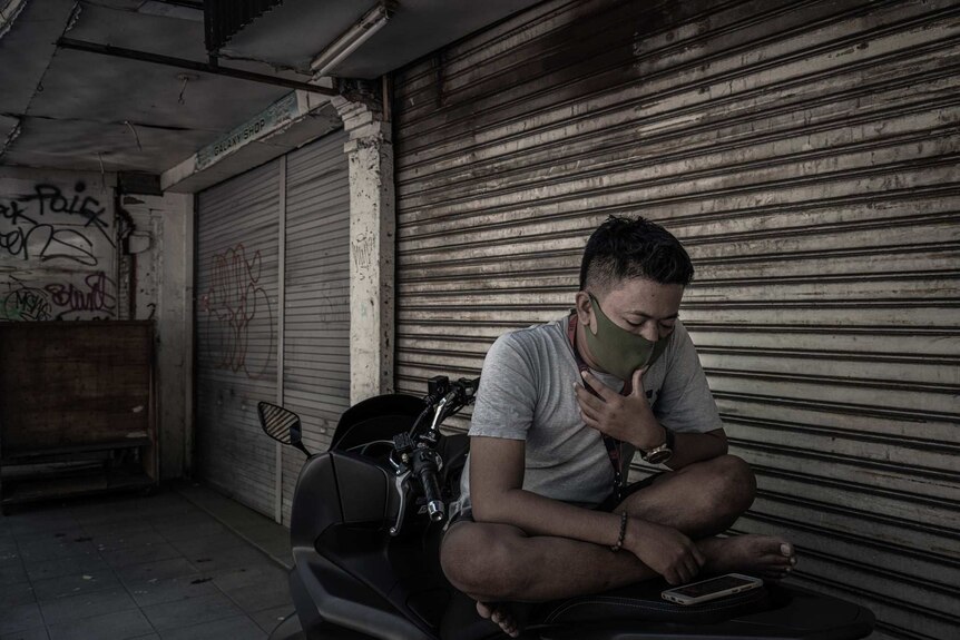 A man wears a mask while sitting on the back of his motorbike in front of closed shopfronts.