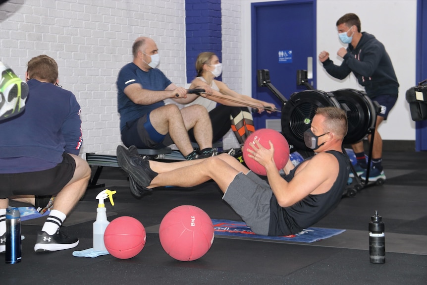 En grupp människor i gymkläder, i ett gym, med en man på golvet med en viktboll och andra som cyklar.