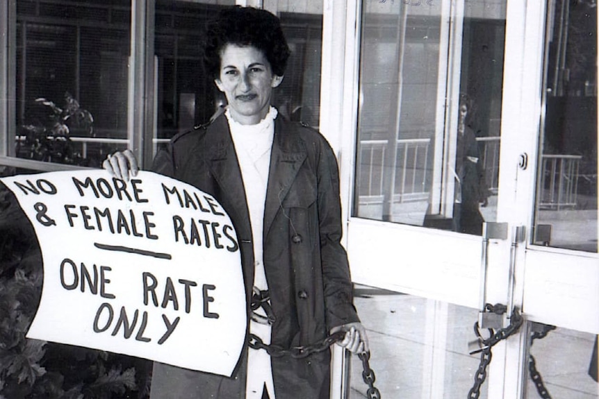 Zelda D'Aprano chained to doors of an office holding a sign that reads "No more male & female rates - one rate only"