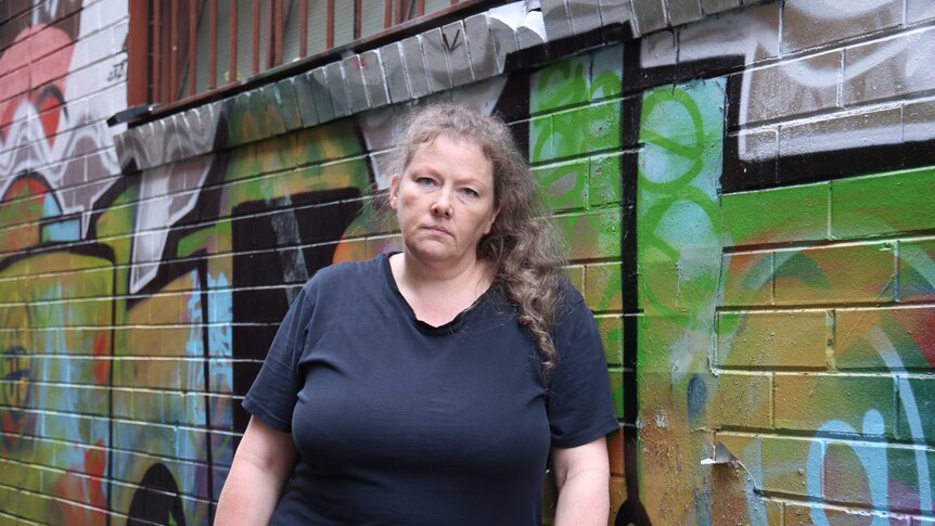 Pamela stands in front of a laneway wall painted with graffiti.
