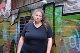 Pamela stands in front of a laneway wall painted with graffiti.