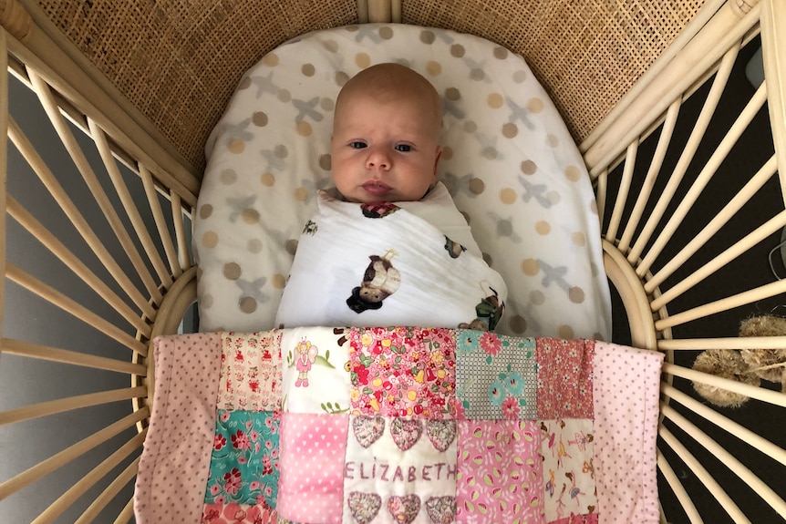 A baby, photographed from above, stares up at a camera, tucked into a pink and green patchwork quilt