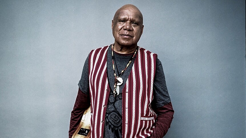 Archie Roach wears a striped vest and a necklace. He is holding a guitar at his side.