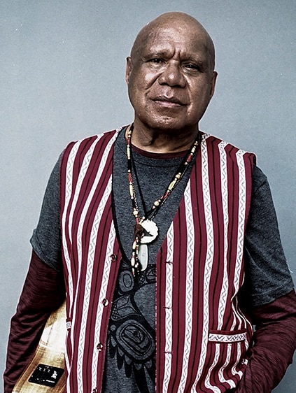 Archie Roach stands wearing a vest holding a guitar