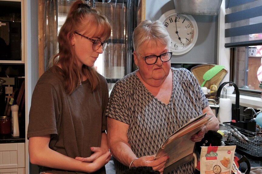 A teenage girl and a woman stand in their kitchen looking at a recipe.