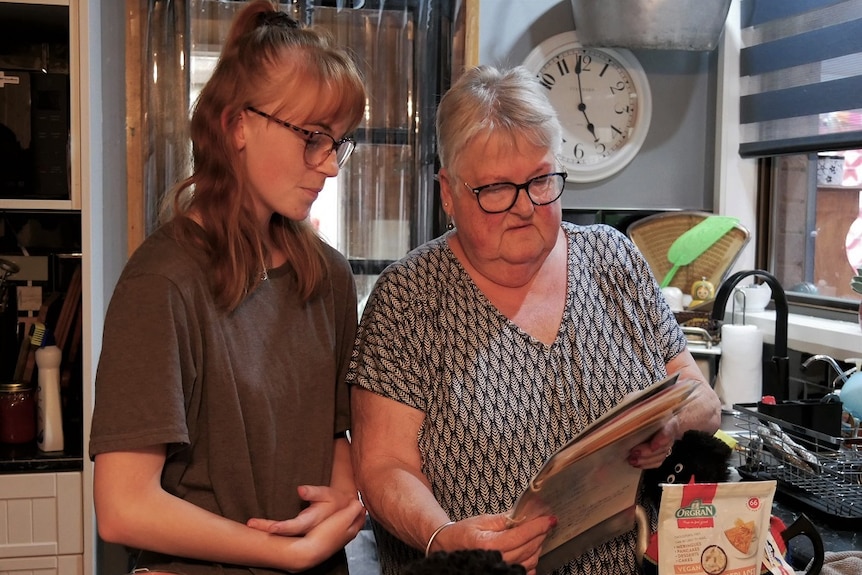 A teenage girl and a woman stand in their kitchen looking at a recipe.