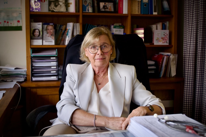 a woman in white jacket and glasses sitting at a work desk in front of a book case