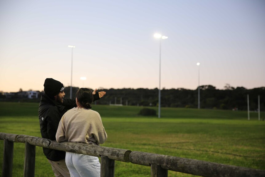 Seen from behind, Trei and Karlie are leaning against a fence in a sports field during twilight.