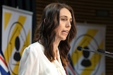 Jacinda Ardern stands at a lectern. Behind her is a New Zealand flag and two signs with yellow and white striped posters