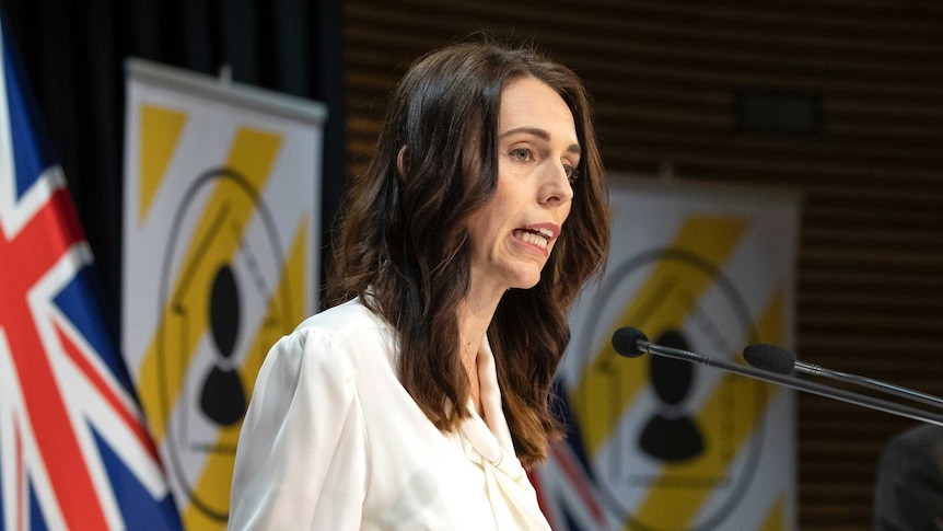 Jacinda Ardern stands at a lectern. Behind her is a New Zealand flag and two signs with yellow and white striped posters