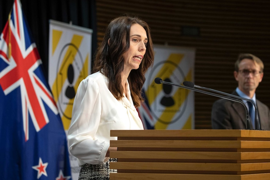 Jacinda Ardern stands at a lectern. Behind her is a New Zealand flag and two signs with yellow and white striped posters