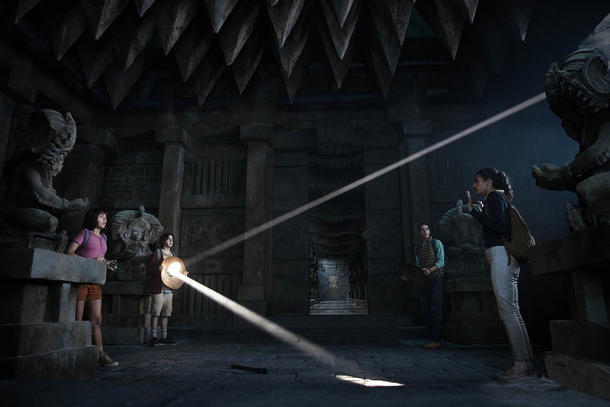 Four teenagers stand in dark ancient site one holding a circular object and reflecting a beam of light.