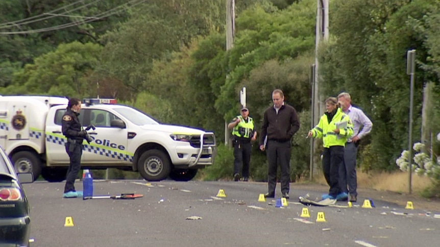 Police stand on a road and point to car debris and police markers.