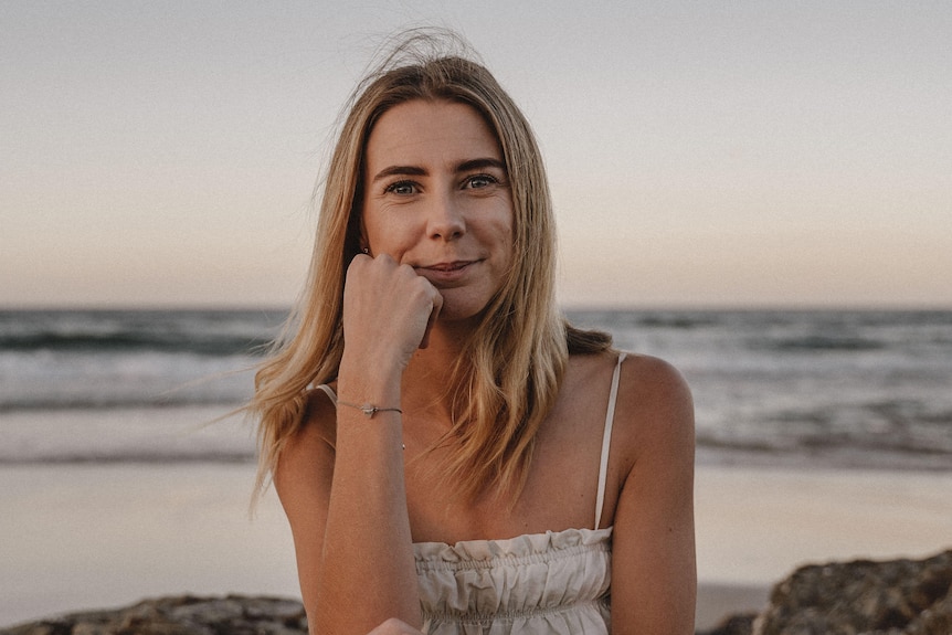 Karla is sitting by the beach at sunset. She is resting her elbow on her knee with her right hand to her face. She is smiling.