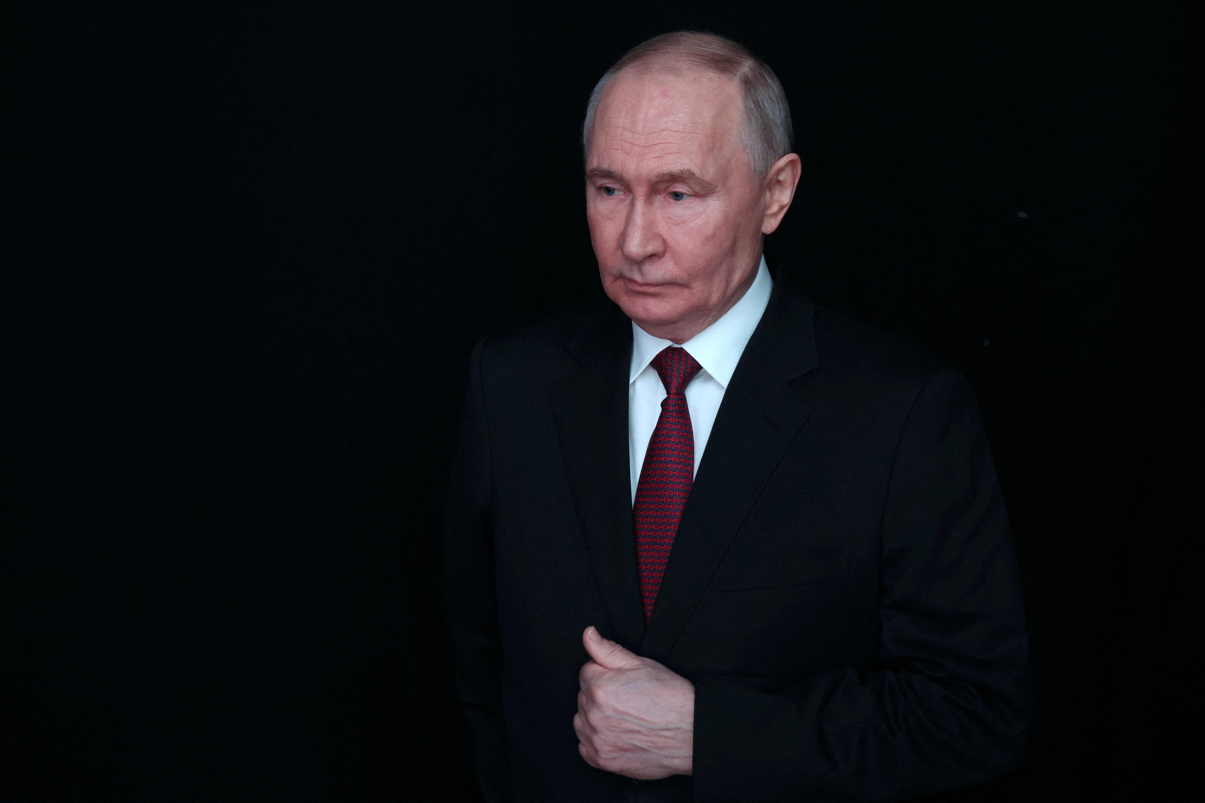 Vladimir Putin wearing a dark suit against a pitch black background.