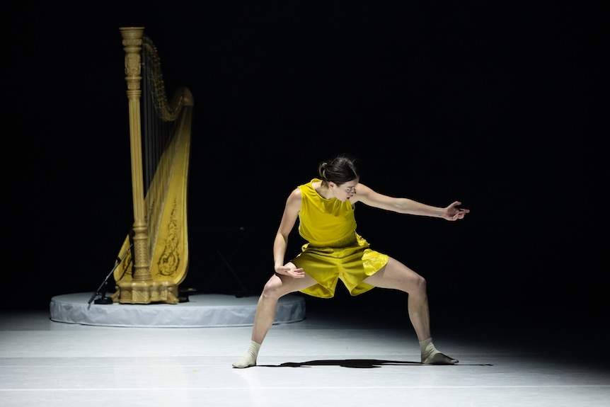 Stage shot showing a solo woman dancer in a yellow tank tops and shorts posing dramatically with a harp in the background. 