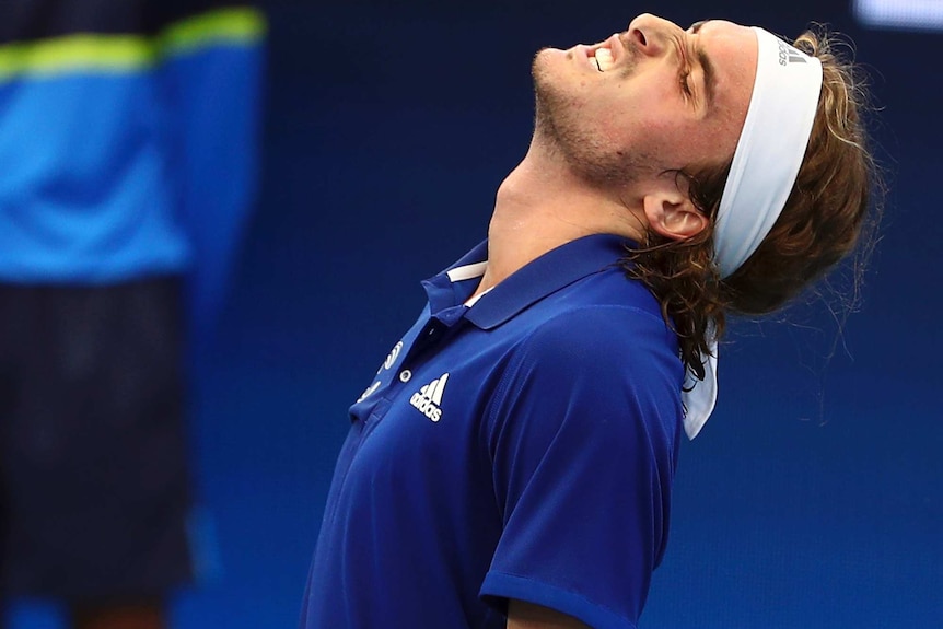 Stefanos Tsitsipas, wearing a blue shirt and shite headband, rocks his head back and grimaces.