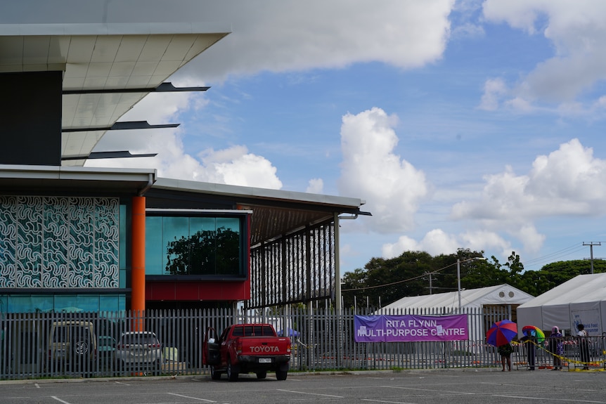 A facility with cars lined up out front with a flag saying Rita Flynn multipurpose centre