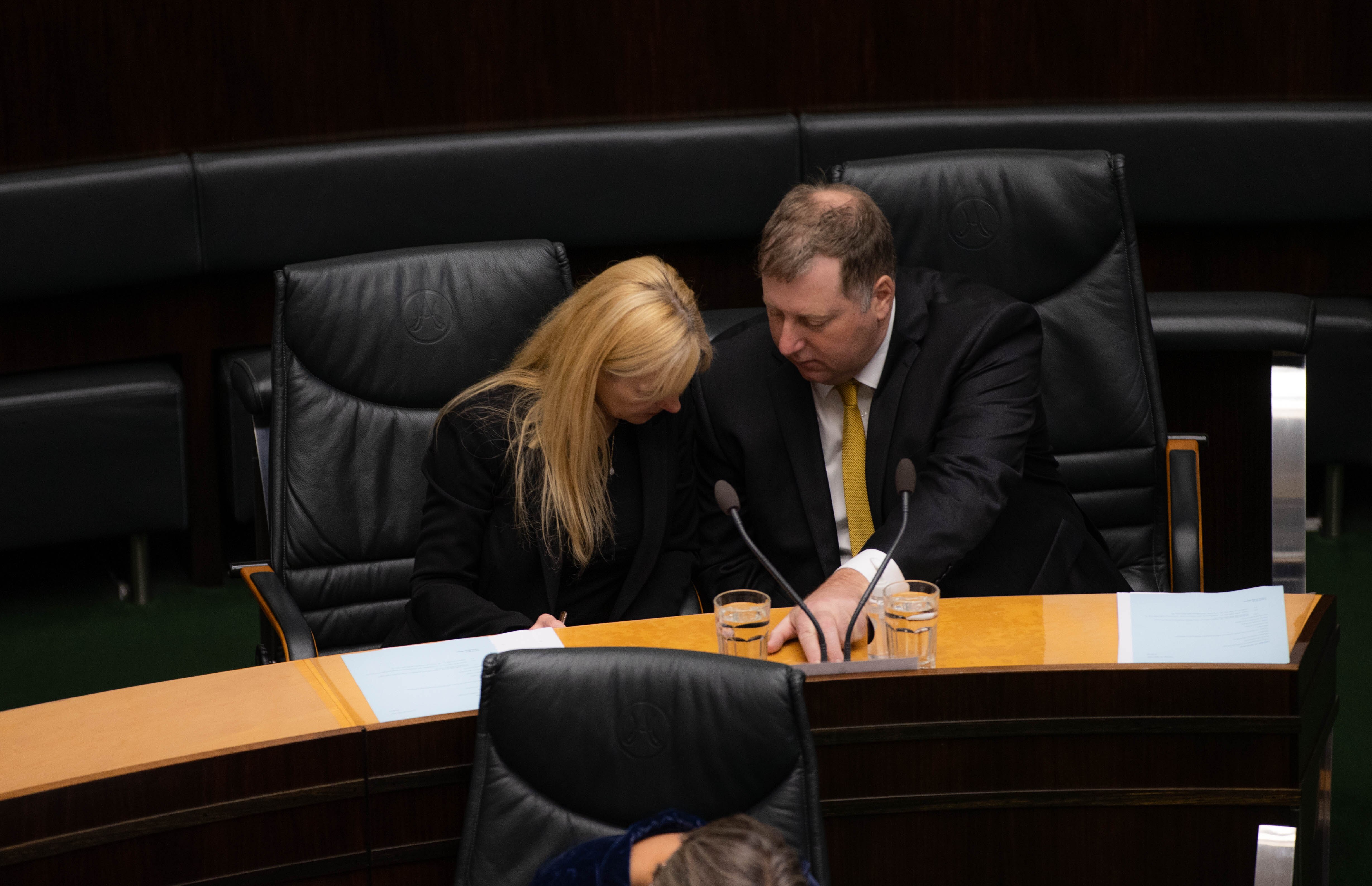 A blonde woman and a man have their heads down looking at documents on a desk in state parliament