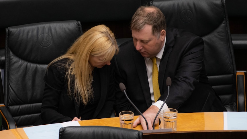 A blonde woman and a man have their heads down looking at documents on a desk in state parliament