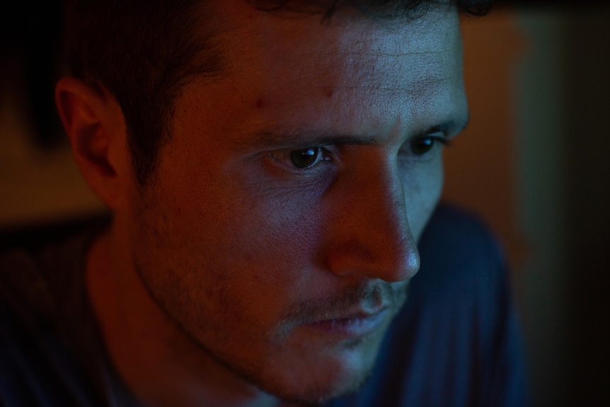 Man looking at a computer screen in a dark room, with red and blue light shining on his face.