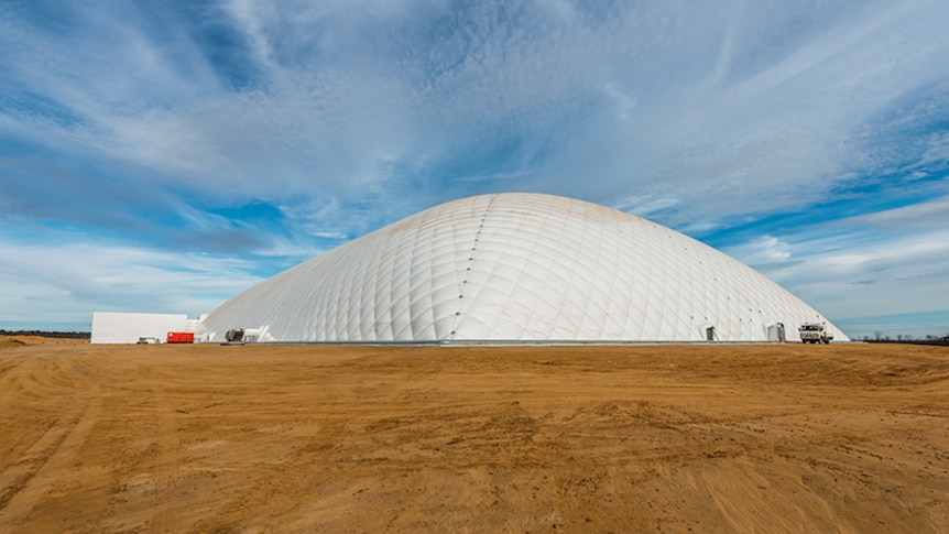 A big white dome rises from the brown dirt landscape under a cloud=streaked blue sky.
