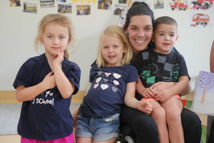 A young woman in a wheelchair cuddles a young boy and two girls, all smiling.