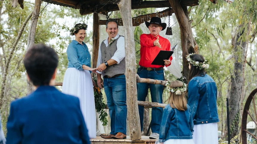 Man dressed as a cowboy wearing a bright red shirt and Akubra-style hat with a couple officiates at a vow renewal ceremony.