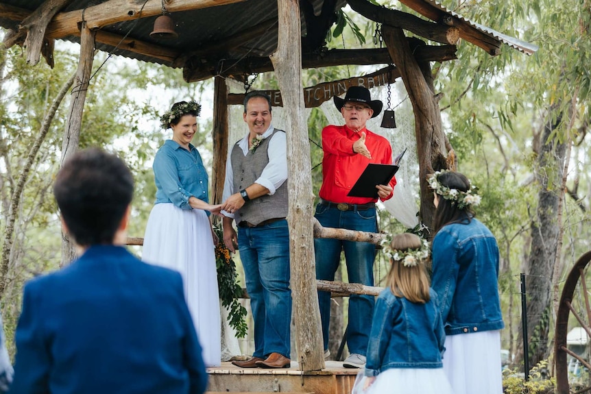 Man dressed as a cowboy wearing a bright red shirt and Akubra-style hat with a couple officiates at a vow renewal ceremony.