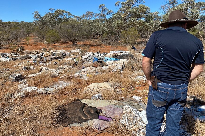 A man is looking to the side with piles of rubbish in front of him, spread throughout brown soil, dry scrub and green trees.