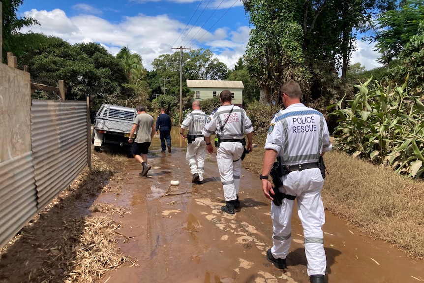 Three police officers in white suits walk down a muddy laneway