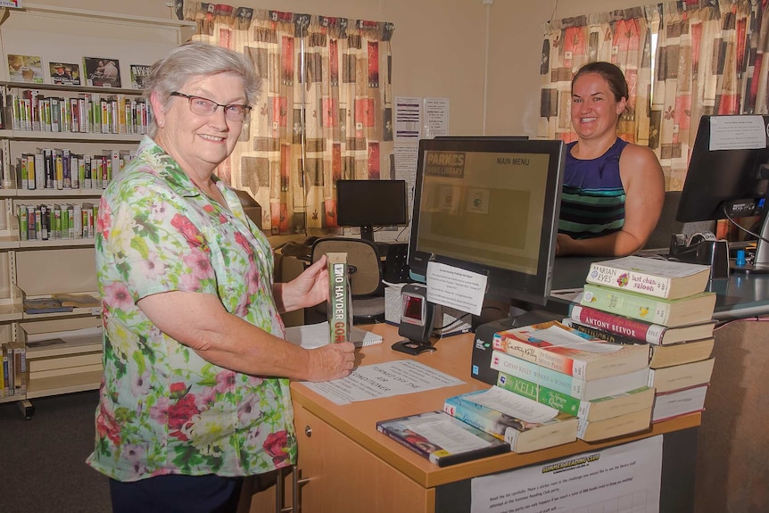 Two women standing in front of computers at a library with shelves of books and audio books in the background.