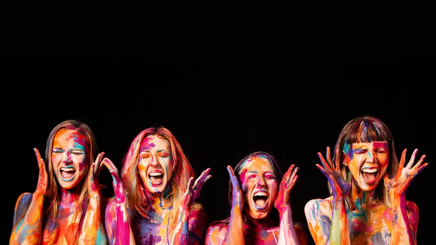 Four women stand in front of a black background. They are covered in body paint and are holding their hands up excitedly.