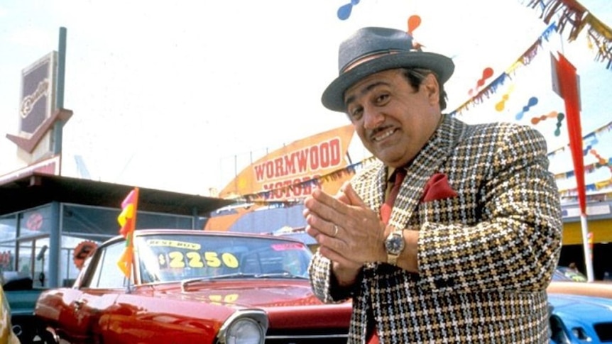 Actor Danny DeVito looks mischievous while standing in front of a used car yard in the film Matilda.