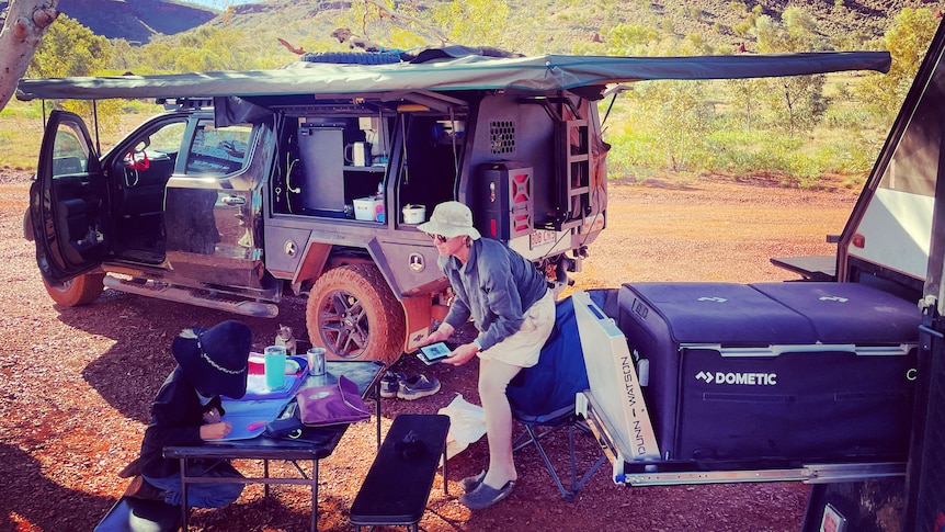 Jo and a child at a plastic camping table and a 4 wheel drive under a gum tree. A treeless mountain range in the background.