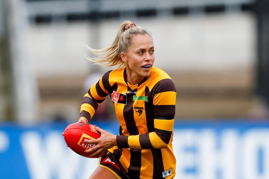 An AFLW player wearing brown and gold stripes, holds the red football in two hands, as she runs